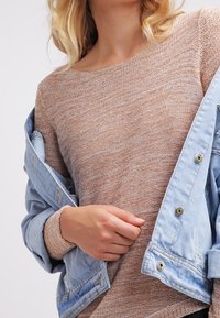 Hellblaue Jeansjacke über einem beigen Strickpullover. Der Pullover hat ein strukturiertes Muster und einen breiten Ausschnitt, mit sichtbaren Bündchen an den Ärmeln.