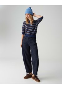 Pull à rayures bleues tricoté à manches courtes, associé à un pantalon large bleu foncé et des baskets marron. Bonnet bleu sur la tête.