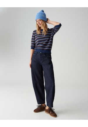 Maglione a righe blu lavorato a maglia con maniche corte, abbinato a pantaloni ampi blu scuro e sneakers marroni. Berretto blu in testa.