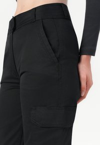 Pantaloni cargo negri, realizați dintr-un material durabil, cu un talie înaltă, buzunare laterale și un buzunar cu clapă pe piciorul stâng.