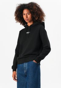 Sweatshirt preto com bolso canguru, capuz com cordão e detalhe do logótipo em branco. Combinado com calças de ganga azuis. Textura de tecido suave.