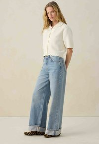 Witte gebreide trui met korte mouwen, gecombineerd met lichtblauwe brede jeans en schoenen met dierenprint, wat zorgt voor een casual uitstraling.