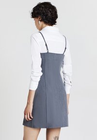 Abito grigio con un tessuto texturizzato, spalline regolabili e un corpetto aderente. Camicia bianca sotto, con maniche arrotolate.