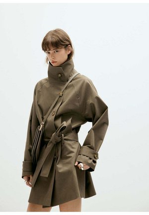 Donna che indossa un trench oversize verde oliva con collo alto, dettagli con bottoni e cintura abbinata, porta una borsa a tracolla, su uno sfondo semplice.