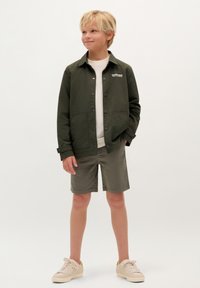 Olijfgroene jas met knoopsluitingen en twee voorzakken, gecombineerd met lichte olijfgroene shorts en beige sneakers. Effen witte t-shirt eronder.