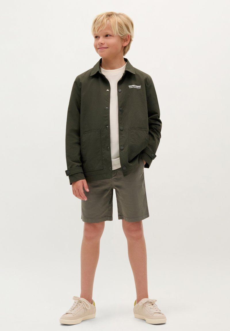 Olijfgroene jas met knoopsluitingen en twee voorzakken, gecombineerd met lichte olijfgroene shorts en beige sneakers. Effen witte t-shirt eronder.