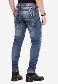 Siniset farkut slim fit -mallilla, joissa on useita takataskuja ja teksturoitua yksityiskohtaa. Mukana erottuva vyötärönauhamuotoilu, jossa on lenkkejä ja yksityiskohtia.