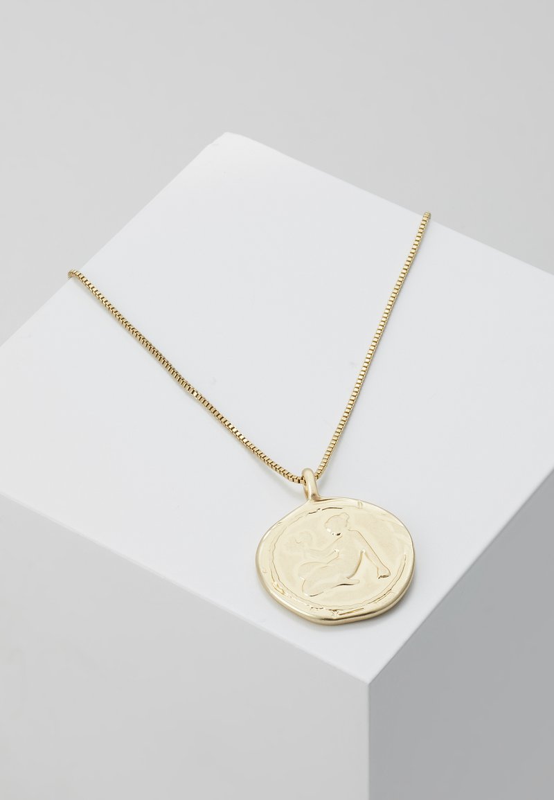 Collar en tono dorado con un colgante redondo de superficie texturizada y diseño de figura débilmente delineado, reposando sobre una caja de exhibición blanca.
