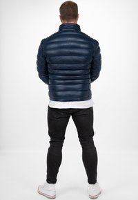 Giacca a vento blu marino, design trapuntato, colletto alto, texture riflettente. Abbinata a jeans neri e sneakers bianche, vista da dietro.