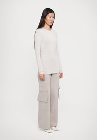 Femme aux longs cheveux bruns portant un haut à manches longues beige clair, un pantalon cargo à jambes larges et des chaussures blanches pointues, se tenant devant un fond blanc.