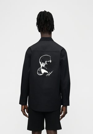 Person mit Dreadlocks trägt ein schwarzes Langarmshirt mit weißer Grafik eines seitlichen Frauenprofils und Unterschrift auf der Rückseite.