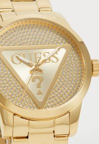 Orologio da polso dorato con quadrante triangolare caratterizzato da una superficie metallica testurizzata e accenti scintillanti. Contrassegnato "GUESS" e un punto interrogativo.
