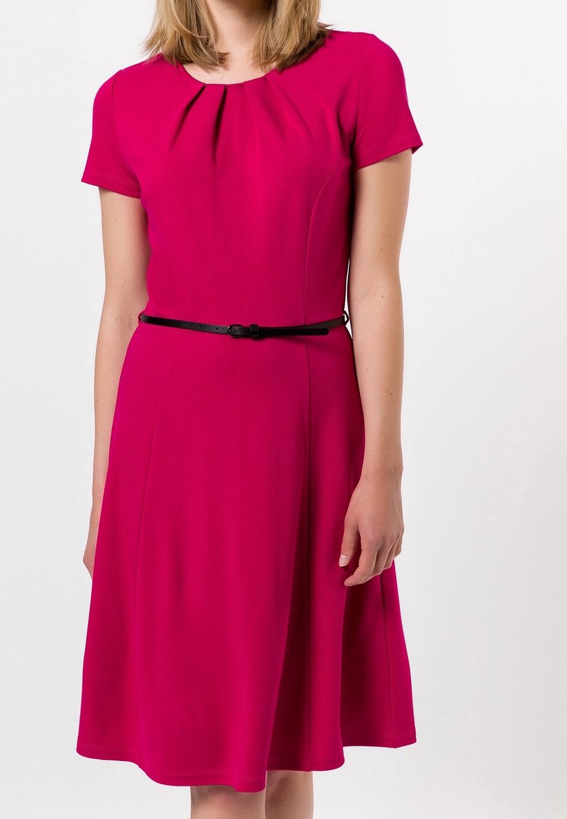 Femme portant une robe rose vif à manches courtes, avec des plis à l'encolure et une fine ceinture noire à la taille.