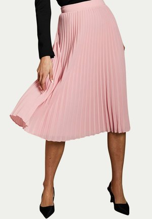 Mujer que lleva una falda midi plisada rosa fluida combinada con mangas largas negras y zapatos de tacón negros puntiagudos.