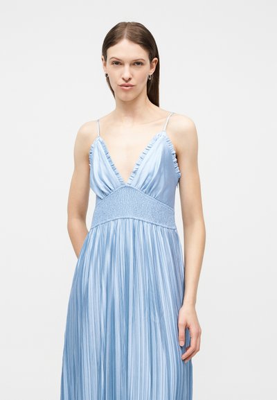 Γυναίκα που φοράει αμάνικο ανοιχτό γαλάζιο φόρεμα με βαθύ V-λαιμόκοψη και βολάν στις άκρες, στέκεται μπροστά σε μια απλή φωτεινή επιφάνεια.