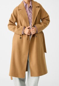 Manteau en mélange de laine camel avec un devant croisé, de grands revers et des poches latérales, porté sur une chemise rayée et un pantalon gris.