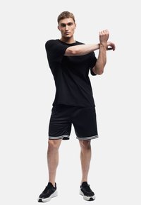 Sort atletisk T-shirt og shorts med hvide stribede detaljer, parret med sorte sneakers. Glat stof og afslappet pasform.