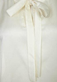 Krémová blůza s hladkým saténovým povrchem, kterou zdobí uvázaná mašle na límečku a dlouhé, ploché stuhy, na jednolitém světlém pozadí.