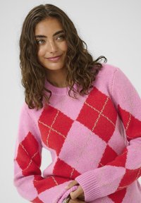 Fiatal nő, göndör barna hajjal, rózsaszín és piros rombuszmintás pulóverben, enyhén mosolyogva és oldalra nézve, sima háttér előtt.