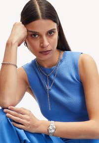 Blauw geribd mouwloos topje, gelaagde zilveren kettingen, zilveren horloge met een gestructureerde band, gladde huid, lang steil donker haar.