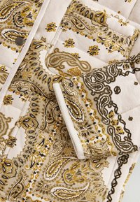 Gequilted stof met een beige basis, voorzien van ingewikkelde gouden en zwarte paisleypatronen. Inclusief een zichtbare pocket en een metalen knoopdetail.