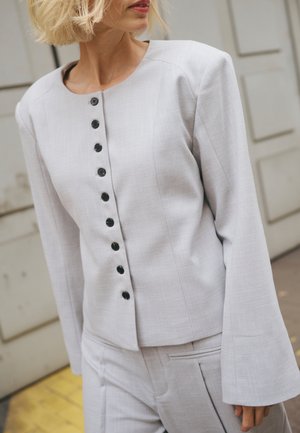 Vrouw draagt een lichtgrijs jasje met knopen en lange mouwen, gecombineerd met bijpassende broek, staand tegen een neutrale achtergrond.