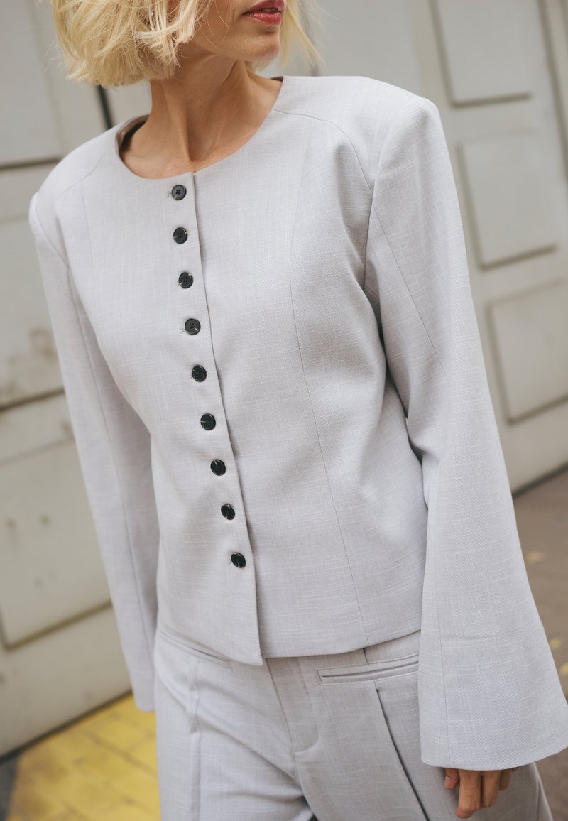 Femme portant un blazer boutonné gris clair à manches longues et un pantalon assorti, debout devant un fond neutre.