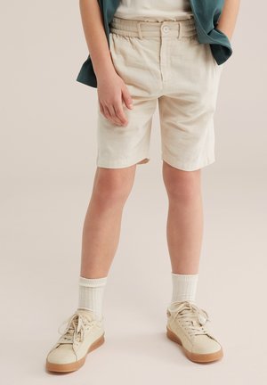 Dziecko ubrane w beżowe szorty, białe skarpetki do kostek i kremowe trampki, stojące z jedną ręką swobodnie opuszczoną wzdłuż ciała.