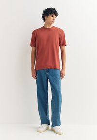 Rozsdabarna rövid ujjú póló kék egyenes szárú farmerral párosítva. A ruházatot fehér sportcipő és minimalista dizájn egészíti ki.