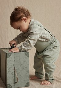 Boîte de rangement avec un motif vert et des accents en métal. Un enfant en chemise à carreaux et en pantalon vert doux jette un coup d'œil par-dessus la boîte, engagé dans le jeu.
