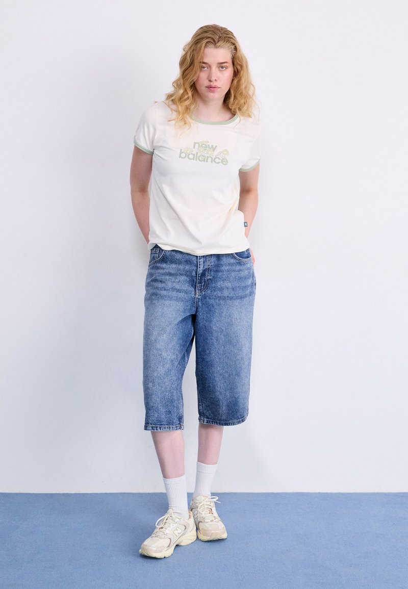 T-shirt bianca con testo verde "New Balance", abbinata a shorts di jeans blu a lunghezza ginocchio e sneakers chiare con calzini bianchi.
