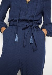 Navy blue textured jumpsuit with a buttoned front, long sleeves, and a tied waist detail featuring tassels.