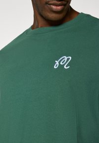 Πράσινο βαμβακερό t-shirt με στρογγυλή λαιμόκοψη και κοντά μανίκια. Ένα λευκό κεντημένο λογότυπο εμφανίζεται στο στήθος. Μαλακή υφή.