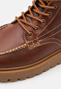 Botte en cuir marron avec des lacets à motifs orange et marron, œillets métalliques, coutures détaillées et semelle en caoutchouc texturée.
