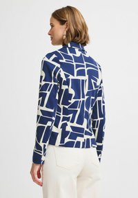Chemise à manches longues bleu marine et blanche, ornée d'un motif géométrique, en tissu doux, avec un col, associée à un pantalon de couleur claire.