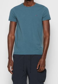 Tyrkysové bavlněné tričko s kulatým výstřihem, krátkými rukávy a malým logem na hrudi. Hladká textura, volný střih.