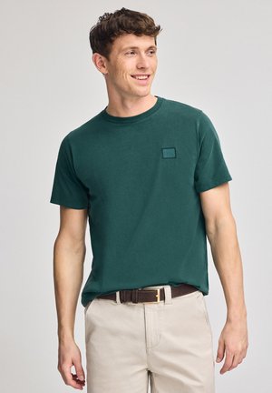 Donkergroene katoenen t-shirt met een ronde hals, korte mouwen en een kleine vierkante logopatch op de linkerborst. Gladde textuur.