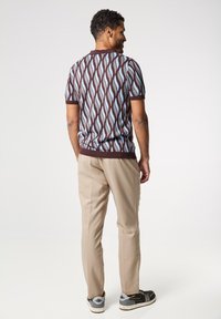 Suéter de manga corta con un patrón marrón y azul, pantalones beige y zapatillas deportivas negras, visto desde atrás sobre un fondo claro.