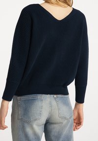 Granatowy dzianinowy sweter o prążkowanej fakturze, z dekoltem w serek i długimi rękawami; noszony z jasnoniebieskimi dżinsami.