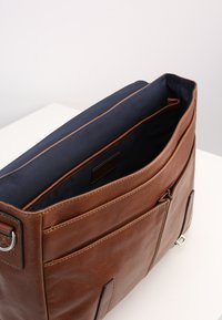 Sac messager en cuir marron avec une surface texturée, doublure marine et plusieurs poches extérieures. Équipé de quincaillerie en argent et d'une fermeture éclair.
