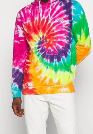 Mann trägt einen mehrfarbigen Batik-Hoodie mit „ellesse“-Logo und weißen Kordeln, kombiniert mit weißen Hosen.