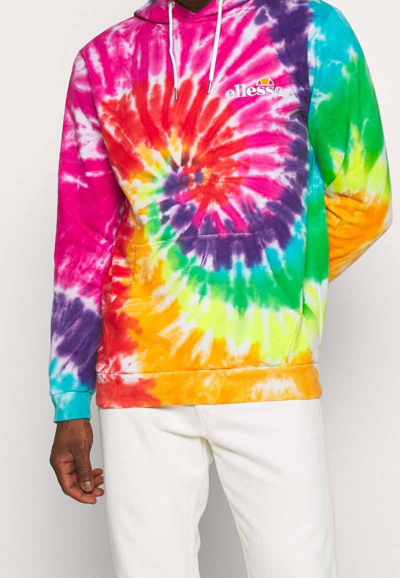 Homme portant un sweat à capuche tie-dye multicolore avec le logo « ellesse » et des cordons blancs, associé à un pantalon blanc.