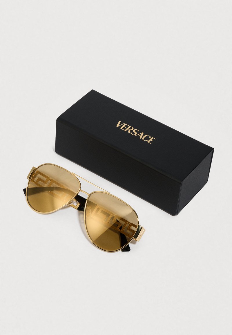 Gafas de sol aviador con filtro dorado, montura de plástico negra y detalles dorados; incluye una caja negra con el logo "VERSACE" en letras doradas.