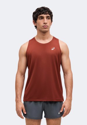 Uomo che indossa una canotta sportiva senza maniche color ruggine e pantaloncini grigio scuro, in piedi davanti a uno sfondo chiaro e semplice.