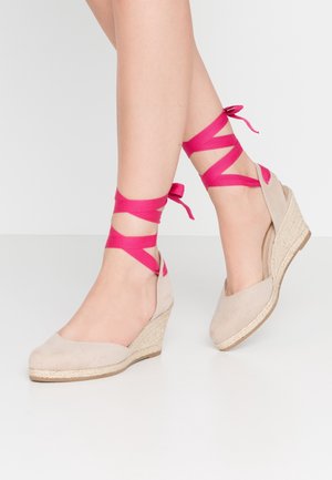 Beige Keilschuhe mit einer strukturierten Oberfläche und einer Jutesohle, ausgestattet mit leuchtend rosa Schleifenbändern, die um den Knöchel gewickelt sind. Glatte, abgerundete Zehenpartie.