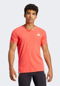 Camisa deportiva de manga corta en coral brillante, con un patrón texturizado, cuello redondo y acentos de logo blanco en el pecho y los costados.