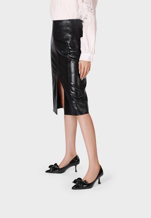 Donna con gonna a tubino nera effetto coccodrillo con spacco frontale, camicetta bianca a manica lunga e décolleté nere con punta appuntita e fiocchi decorativi.