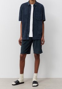 Mörkblå terryklädes kortärmad skjorta med en ficka, i kombination med matchande shorts. Bärs med svarta tofflor och krämfärgade strumpor.