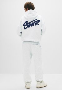 Grijze hoodie met een marineblauw logo op de achterkant, gecombineerd met bijpassende grijze joggingbroek. Zachte textuur en een ontspannen pasvorm. Witte sneakers.
