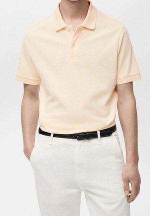 Persona vistiendo una camisa polo de manga corta de color melocotón claro, metida en unos pantalones blancos con un cinturón negro, de pie frente a un fondo liso.